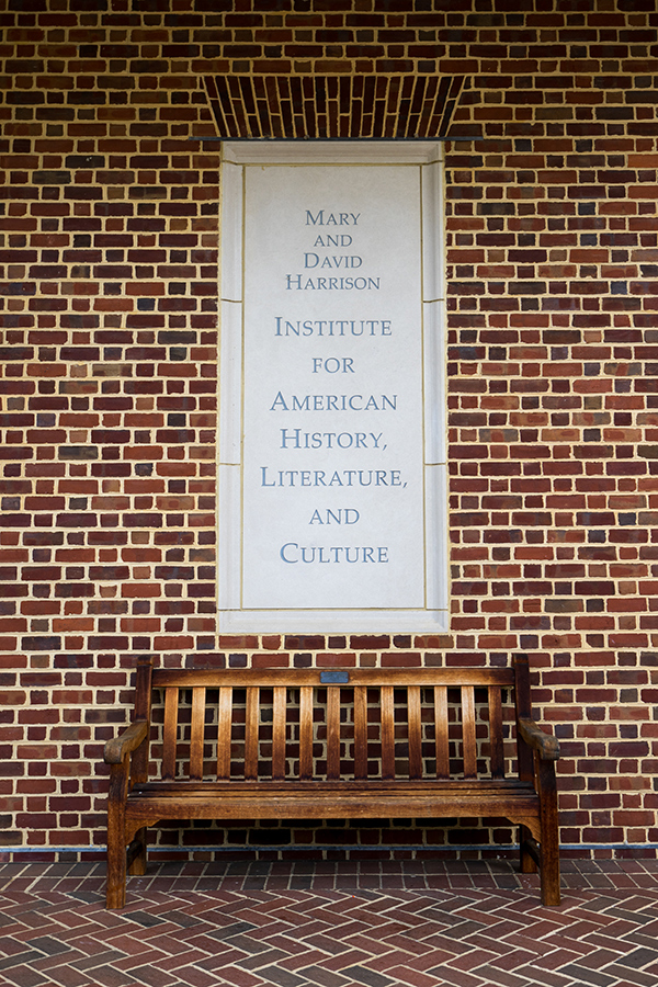 Outdoor plaque with name of building on brick wall, wooden bench underneath.