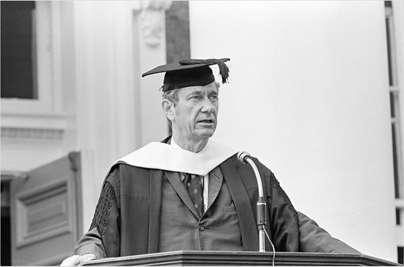 A black and white photo of a white, middle-aged man (Edgar Shannon) in academic robes standing at a podium and speaking into a microphone.