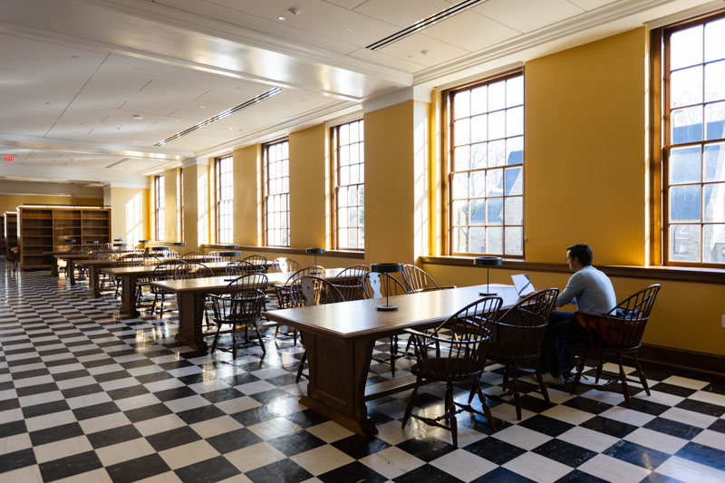 A student sits at a long table working in a large room with yellow walls, big windows, a checkered floor, and bookshelves.